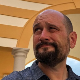 A man with beard staring off camera while standing under an elaborate archway.